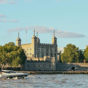 View of the historic Tower of London with a boat on the River Thames under a clear blue sky.