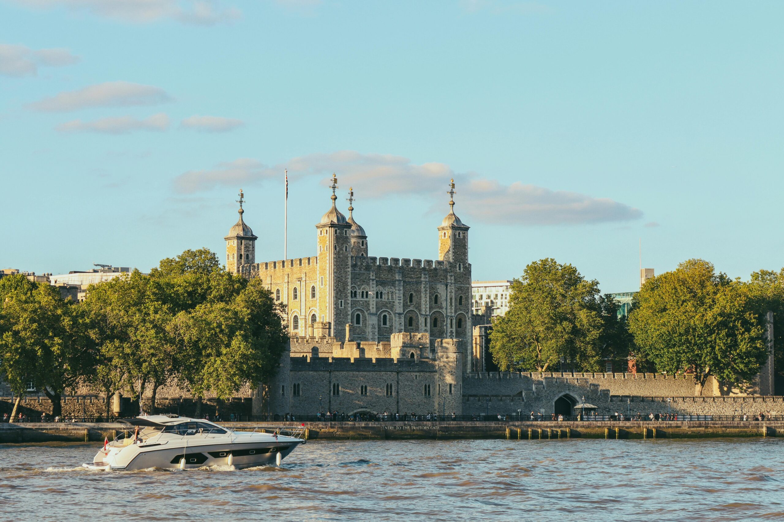 View of the historic Tower of London with a boat on the River Thames under a clear blue sky.