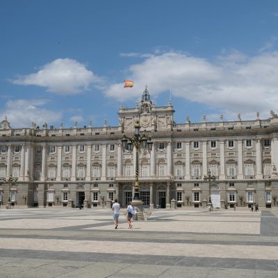 Madrid Royal palace A wide view of the Royal Palace of Madrid with people and the Spanish flag under a blue sky.